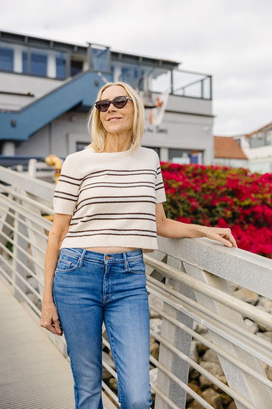 A woman wearing a cream short-sleeve knit top with thin brown horizontal stripes and blue jeans, standing on an outdoor walkway with a railing and buildings in the background.