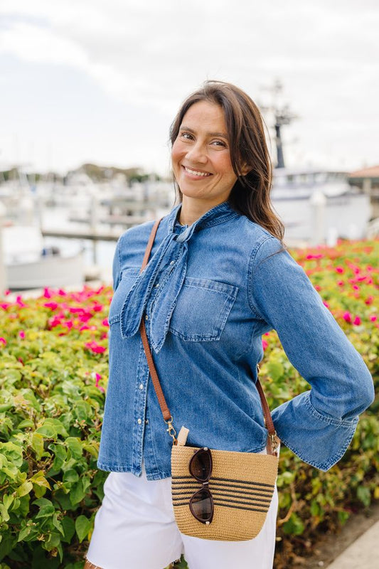 A woman wearing a blue denim shirt with a tie-neck detail and white shorts, carrying a woven straw crossbody bag with sunglasses, standing outdoors in front of green foliage and pink flowers.