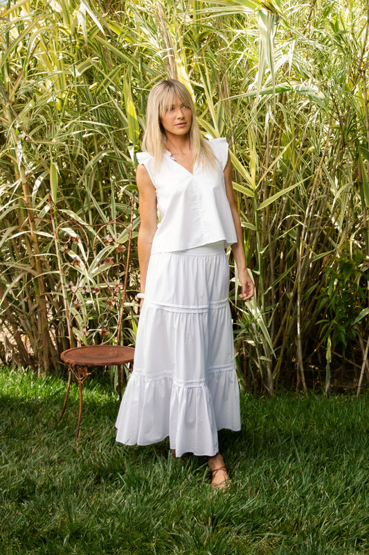 Woman in a white skirt and top standing in a grassy area with tall plants.